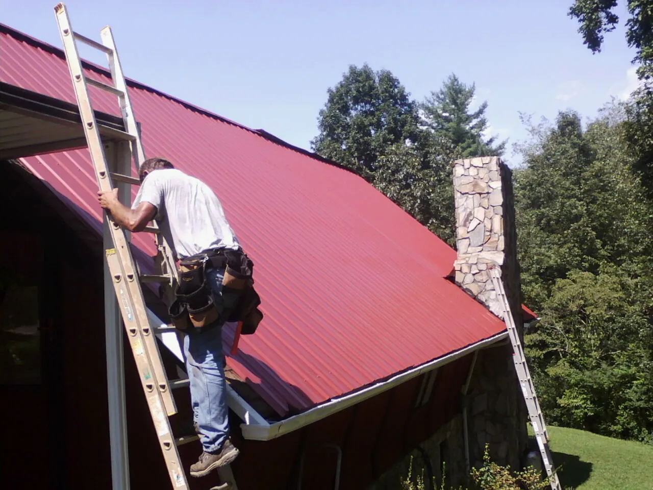 Roofer on a ladder beside a red metal roof and stone chimney — Soffit & Fascia Repair by Roofing Brothers in Prospect Park
